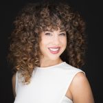 Woman with curly hair smiling, wearing a white sleeveless top, against a dark background.