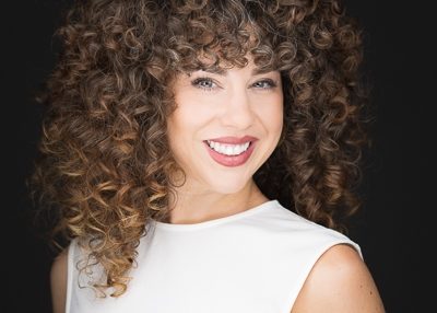 Woman with curly hair smiling, wearing a white sleeveless top, against a dark background.