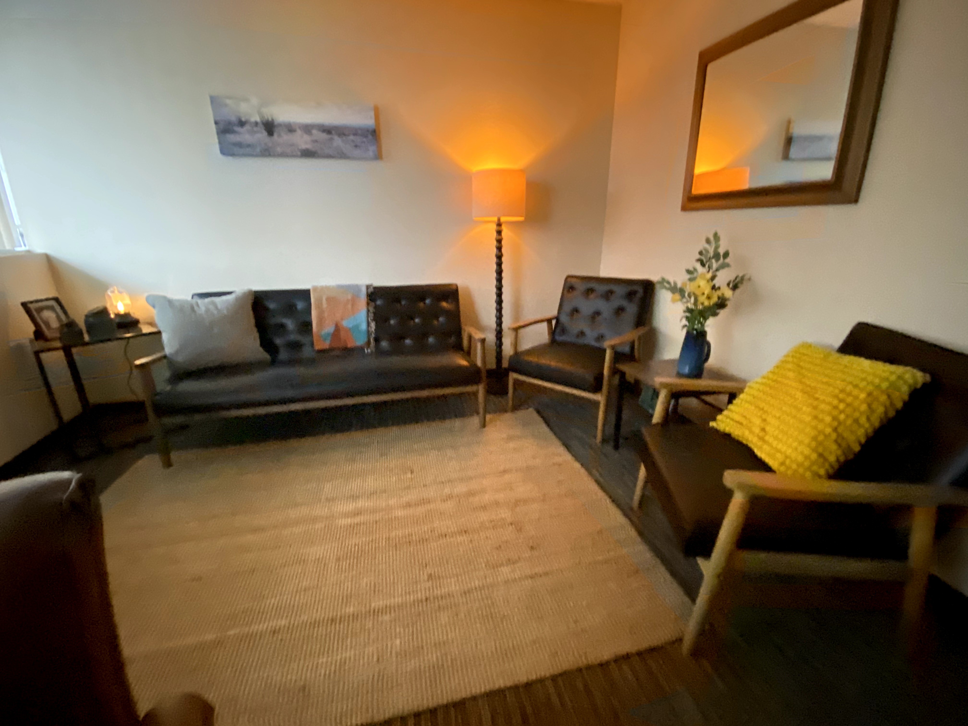 Living room with two black sofas, a wooden chair, a floor lamp, a mirror, and a rug, with a small table and flowers.