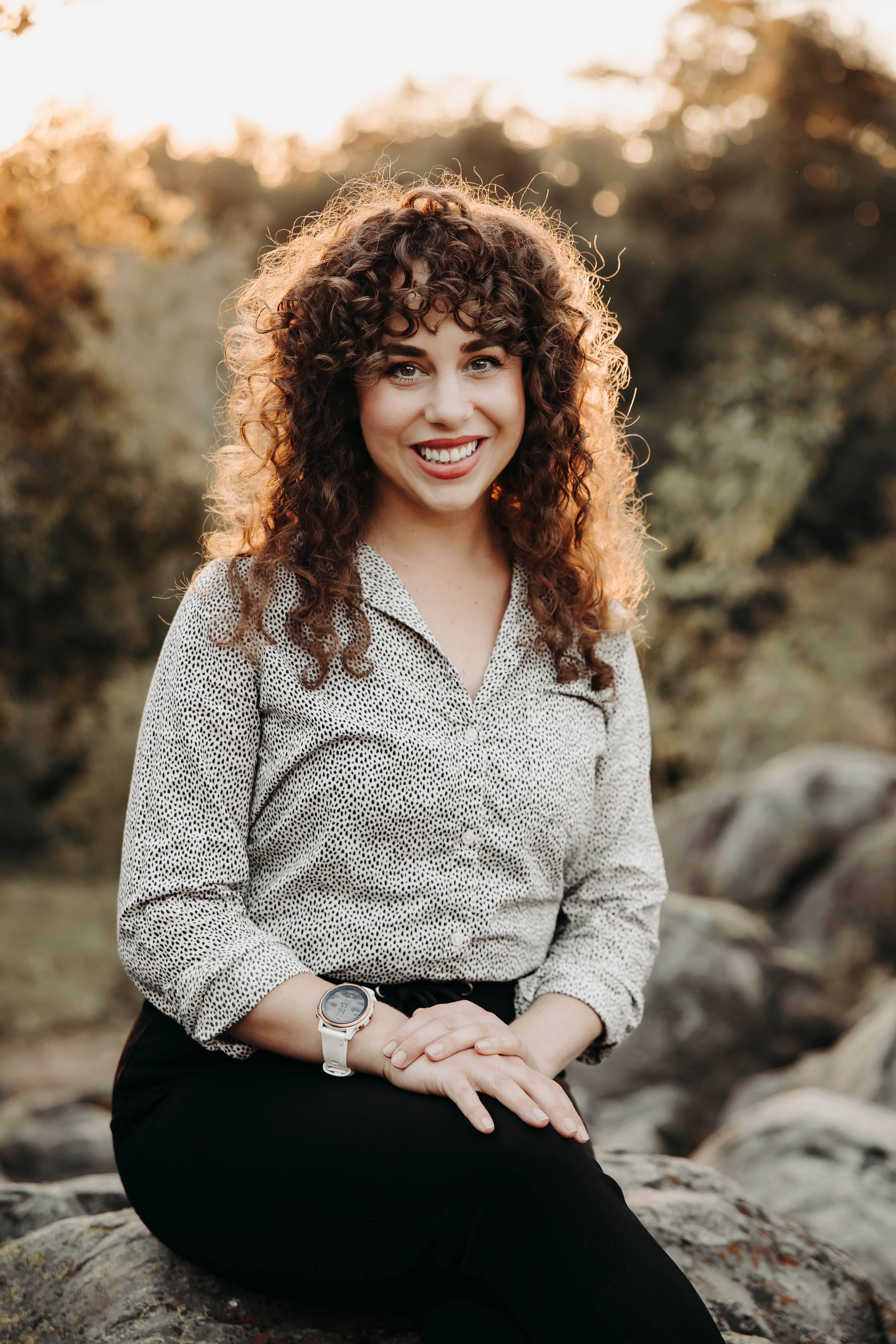Young woman with curly hair smiling outdoors, wearing a patterned blouse and black pants, sitting on rocks.