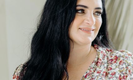 Woman with long dark hair smiling, wearing a floral top, standing indoors near a plant.