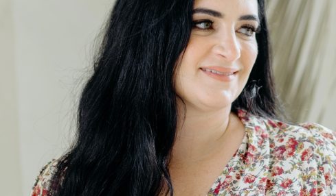Woman with long dark hair smiling, wearing a floral top, standing indoors near a plant.