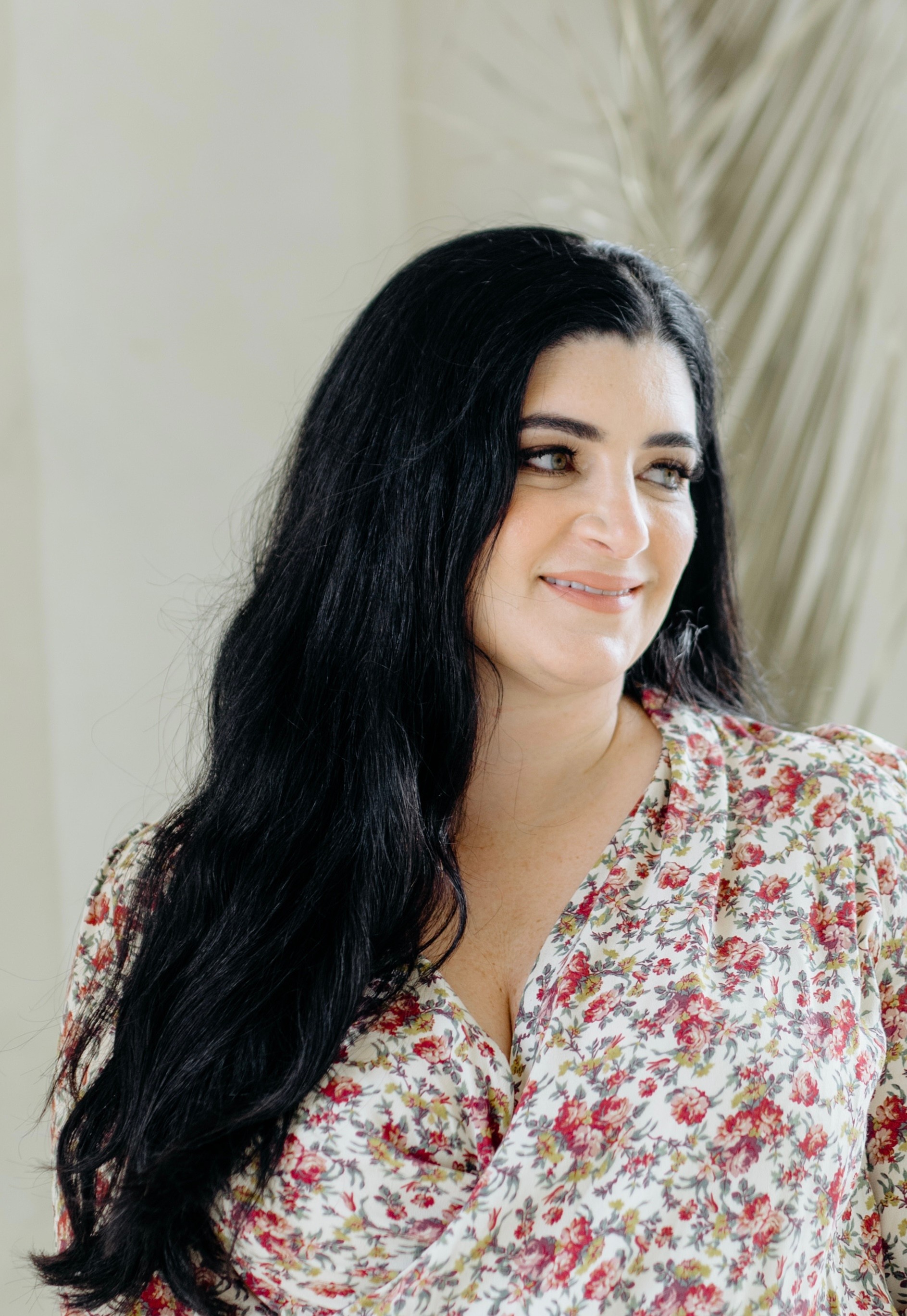 Woman with long dark hair smiling, wearing a floral top, standing indoors near a plant.