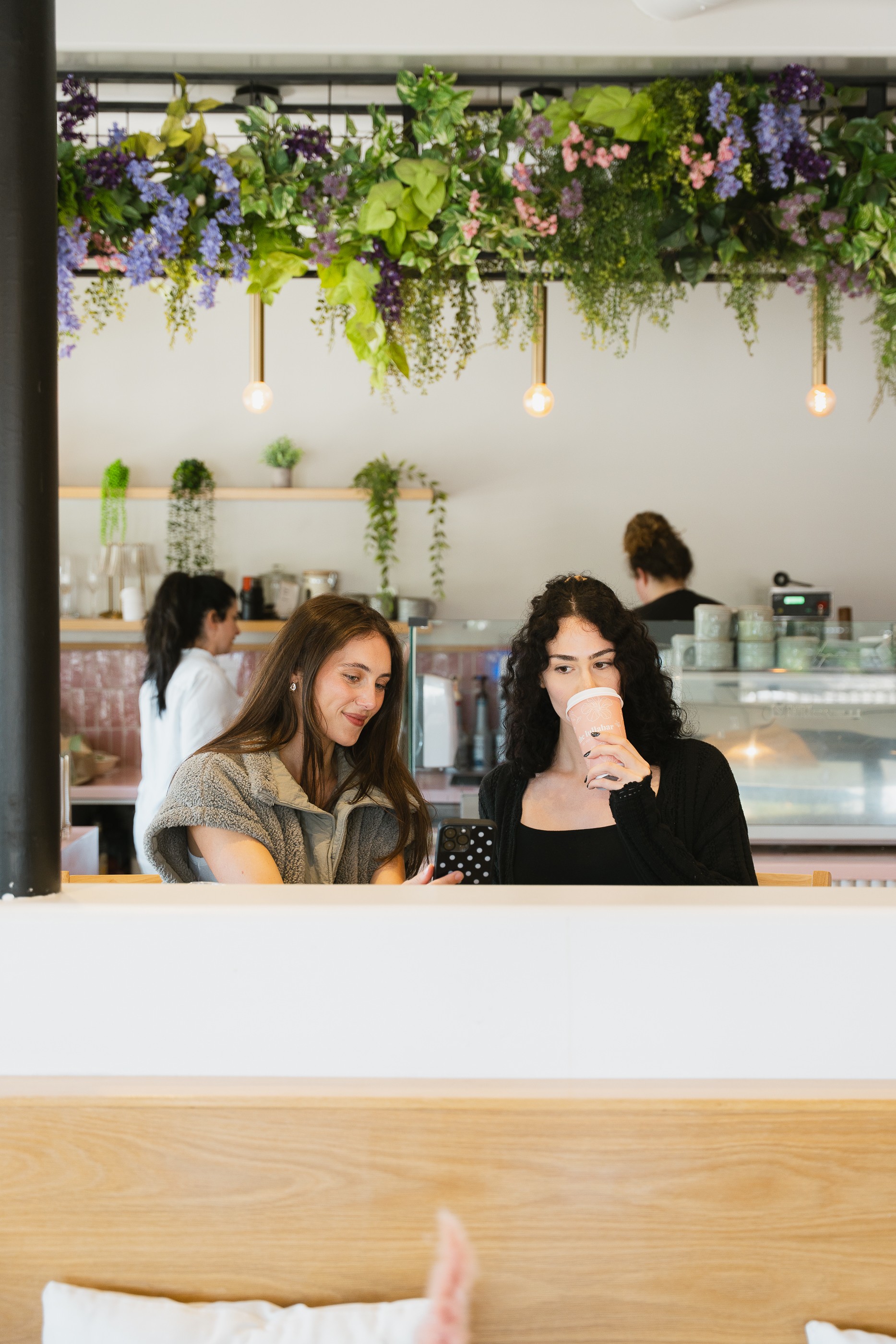 Two women sit at a table in a cafe, one with long hair and the other with curly hair, with hanging flowers above.