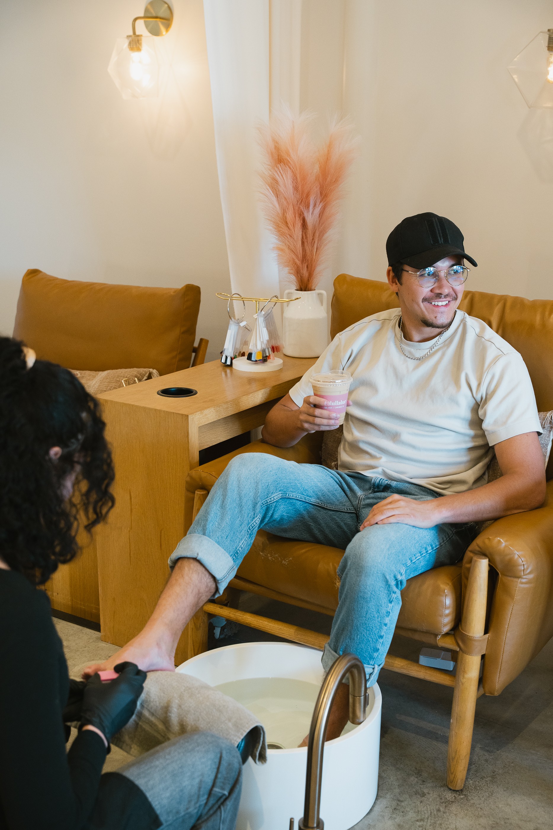Young man with glasses and a cap sitting on a yellow armchair, holding a drink, smiling, with a woman in the foreground. Word count: 20