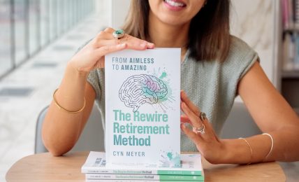 Smiling woman sitting at a table with a book titled 'The Rewire Retirement Method' in front of her.