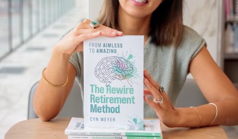 Smiling woman sitting at a table with a book titled 'The Rewire Retirement Method' in front of her.