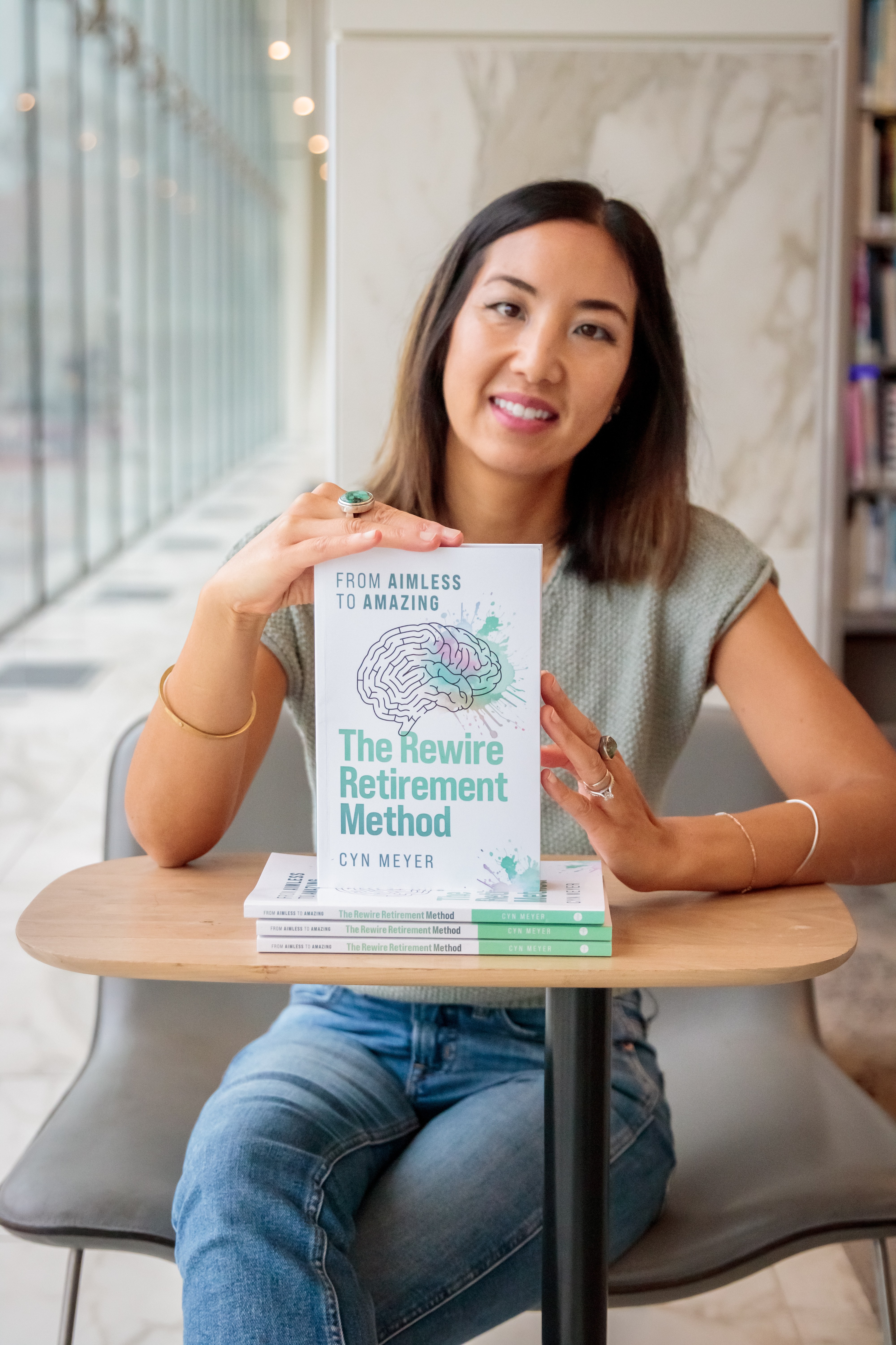 Smiling woman sitting at a table with a book titled 'The Rewire Retirement Method' in front of her.