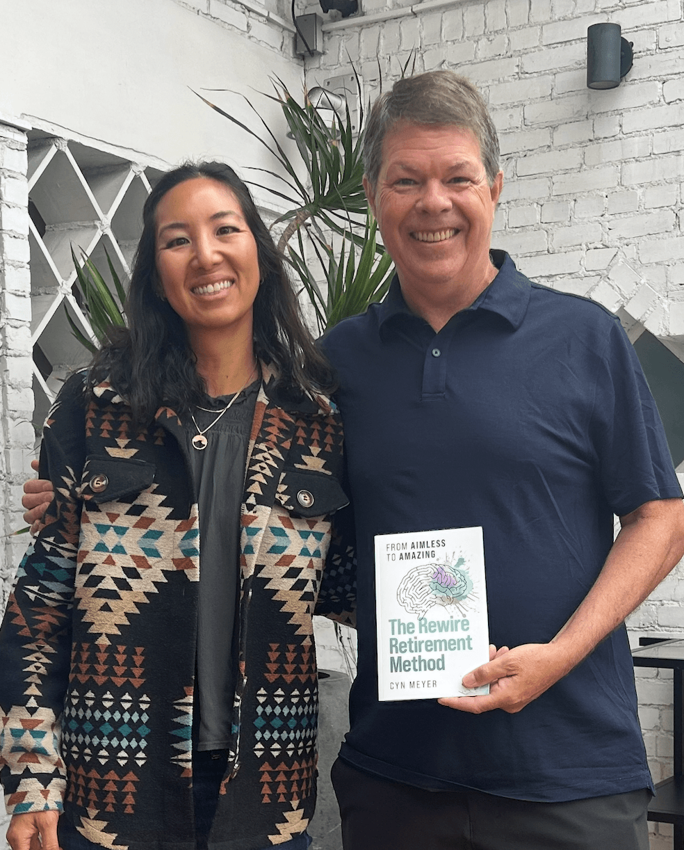 Two people smiling, one holding a book, standing outdoors with a white brick wall and plants in background.