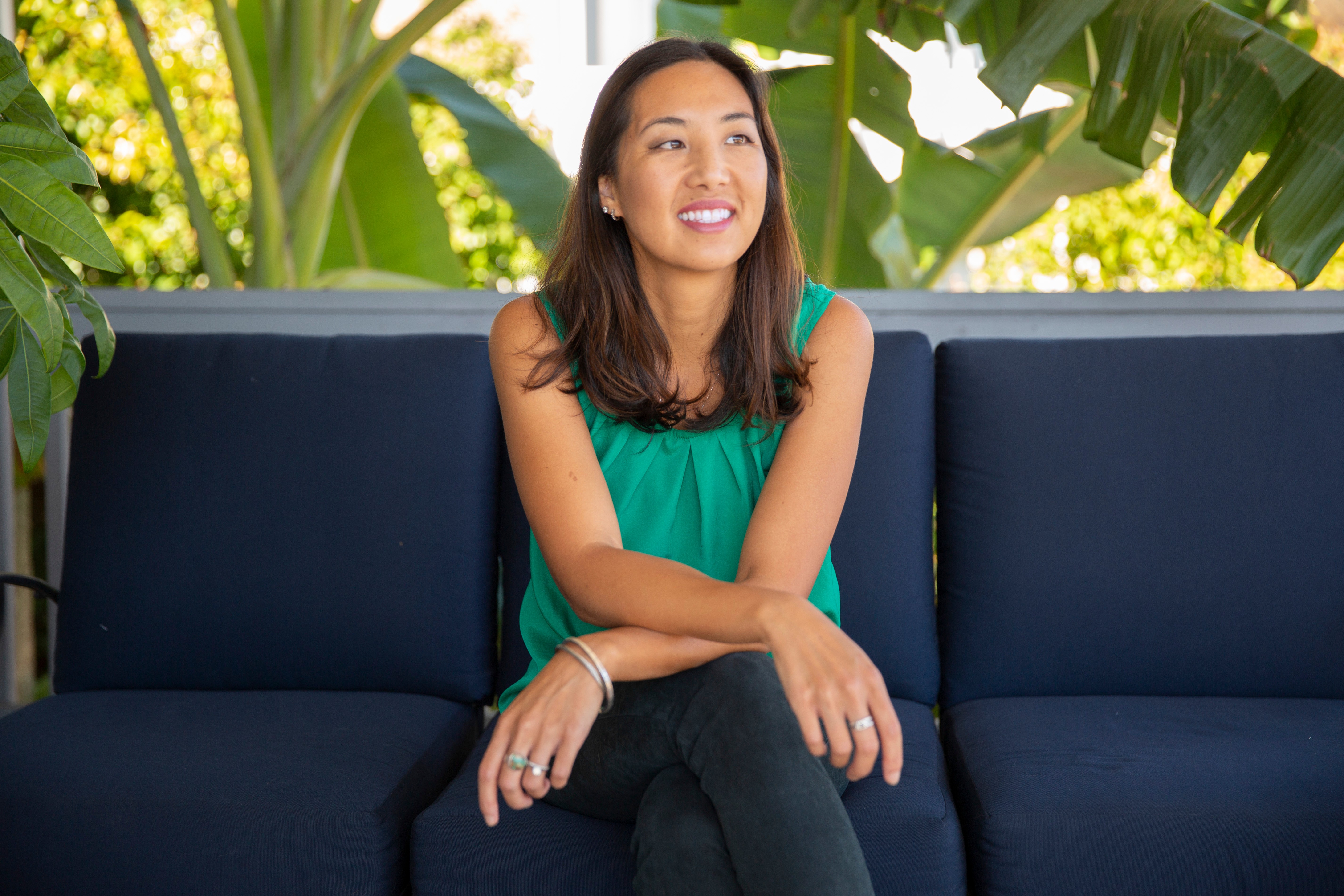 Woman sitting on a dark blue sofa outdoors, smiling, with green plants in the background.