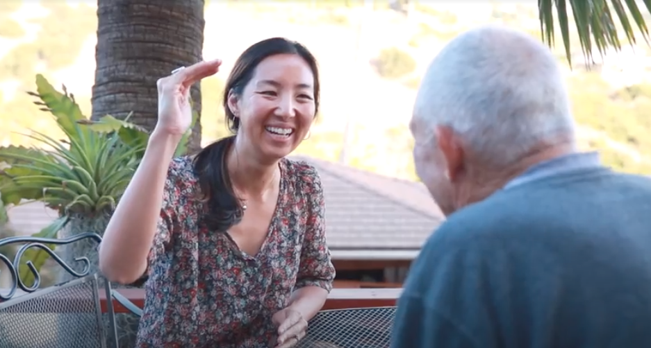 Two people talking outdoors, one woman smiling and gesturing, the other person with gray hair seen from behind.