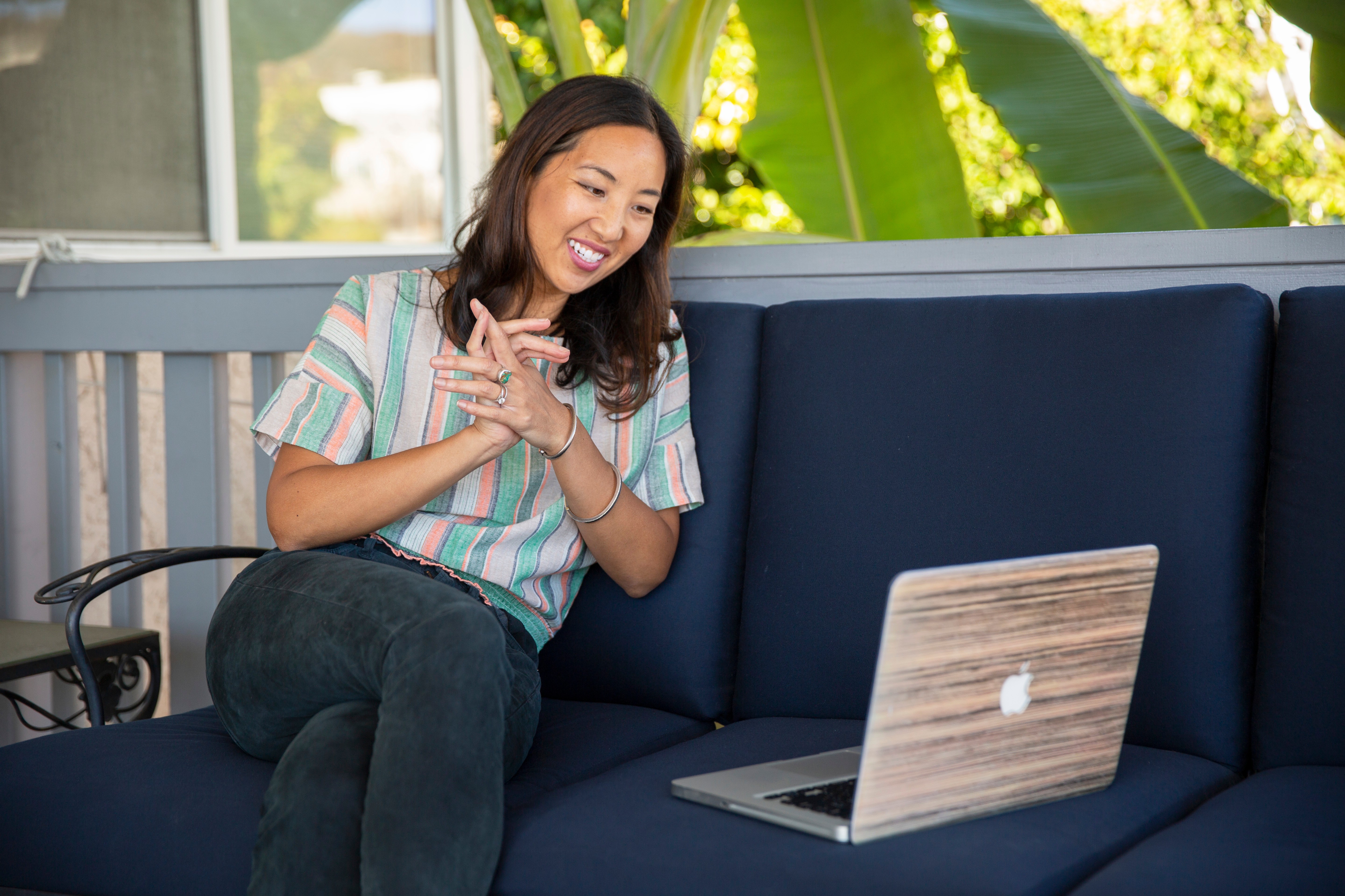Woman smiling and clapping while sitting on a blue outdoor sofa with a laptop nearby.