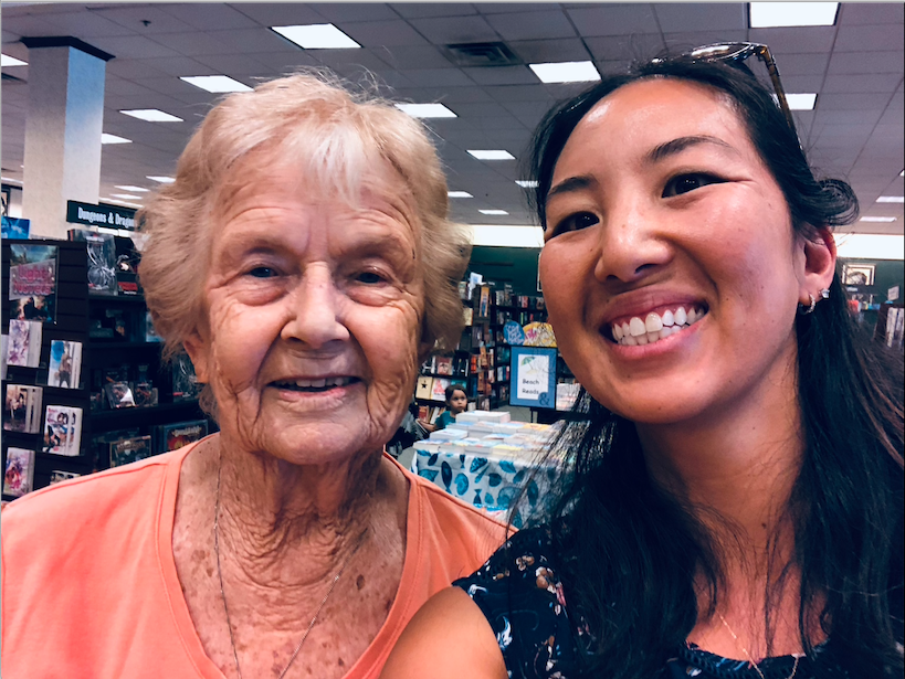 Two women smiling, one elderly with short light hair, the other younger with long dark hair, in an indoor setting.