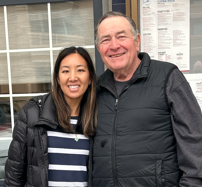 Two smiling people, a woman and a man, standing close together outdoors in front of a window and a menu board.
