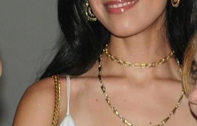 Woman with black hair wearing a white dress, gold jewelry, and smiling.