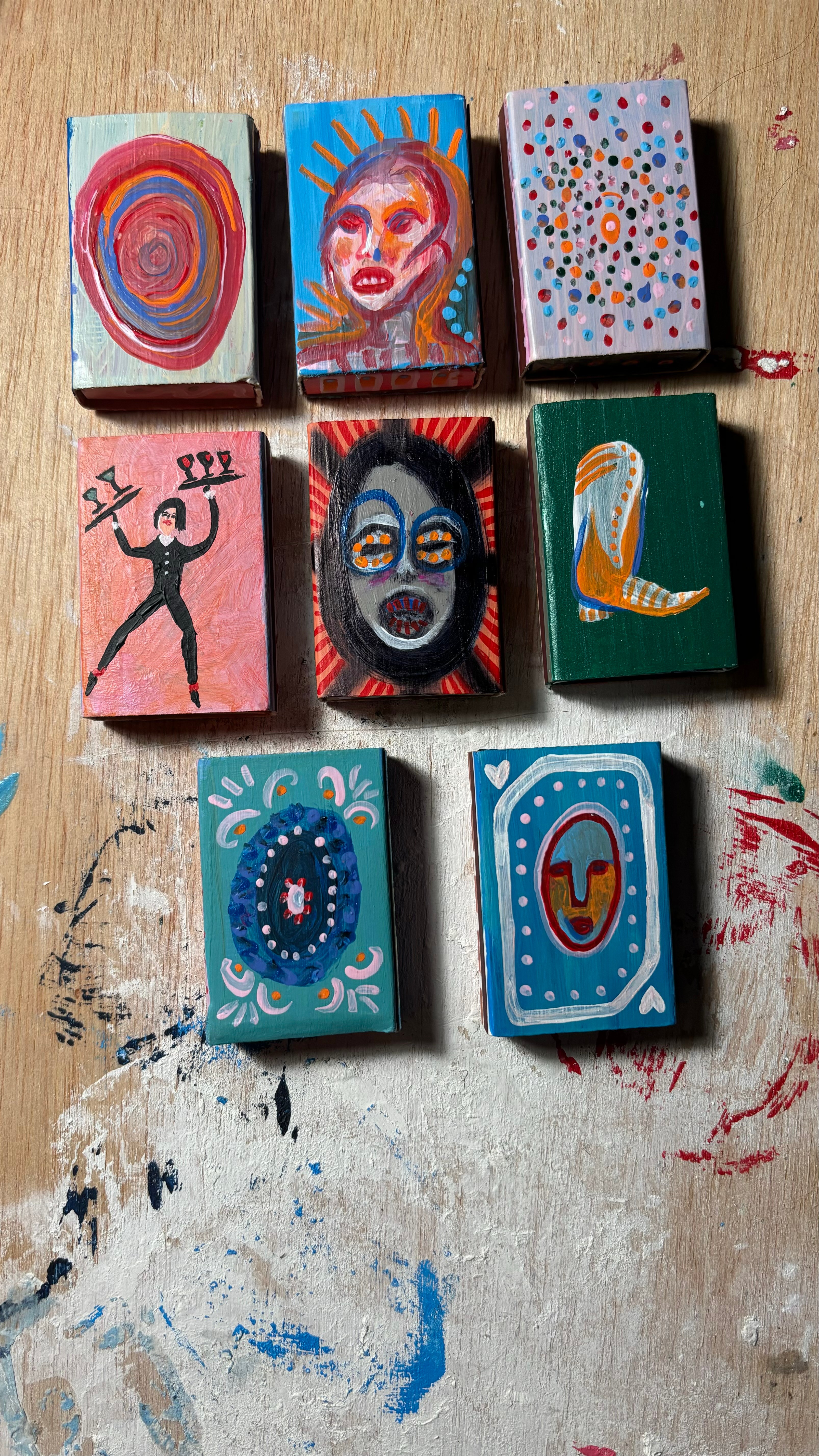 Nine small painted boxes arranged in three rows on a wooden surface, featuring abstract and face designs.