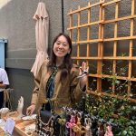 Smiling woman standing outdoors near a wooden trellis with hanging keychains, holding up her hand in a wave.
