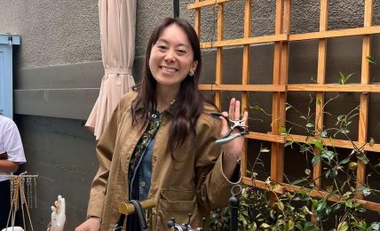 Smiling woman standing outdoors near a wooden trellis with hanging keychains, holding up her hand in a wave.