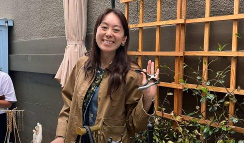 Smiling woman standing outdoors near a wooden trellis with hanging keychains, holding up her hand in a wave.