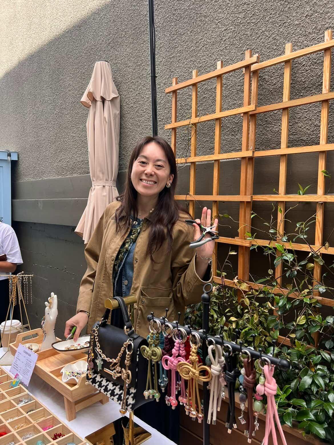 Smiling woman standing outdoors near a wooden trellis with hanging keychains, holding up her hand in a wave.