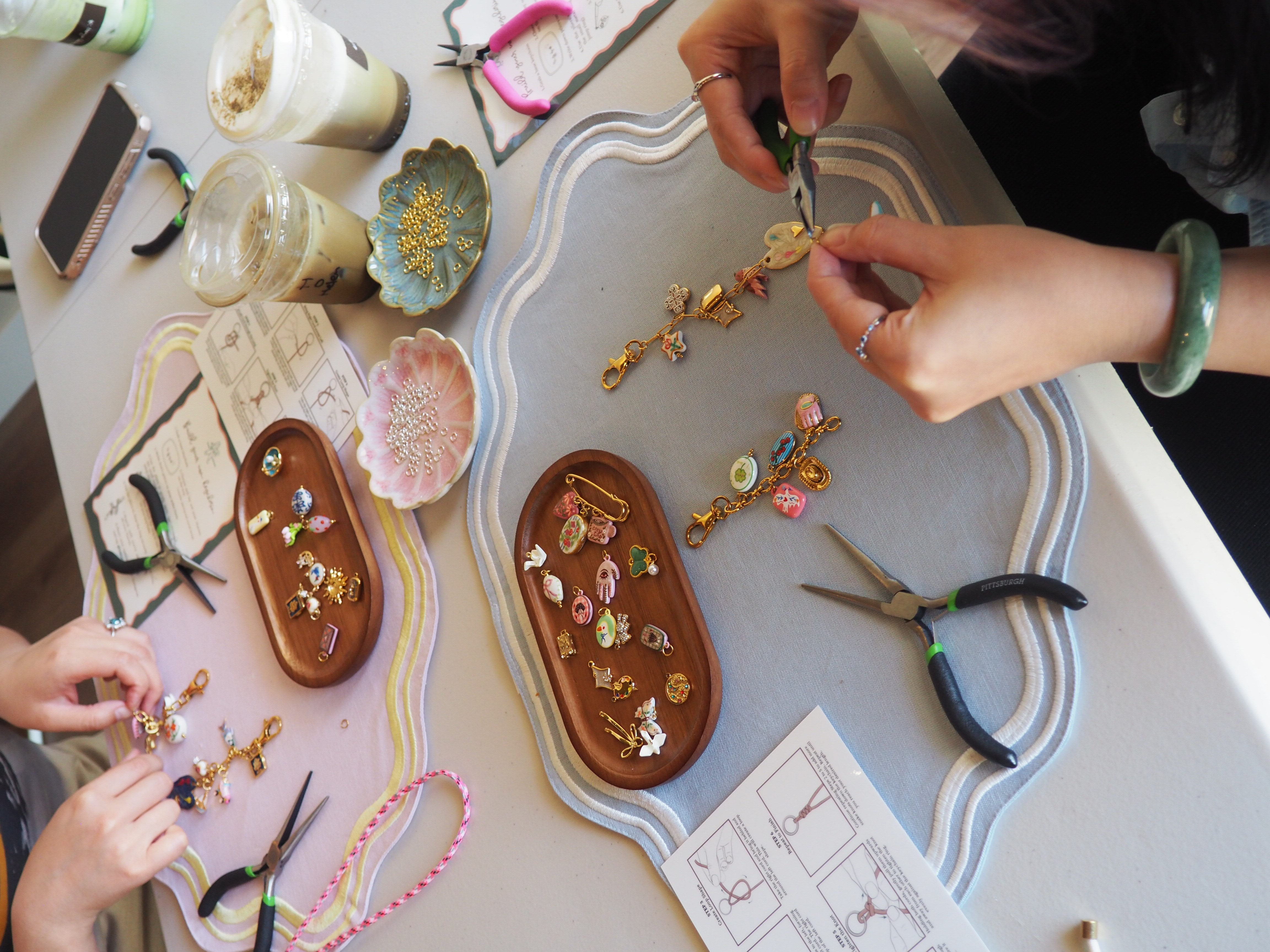 Hands working on jewelry with pliers, beads, and jewelry tools on a table, with jewelry pieces and supplies around.