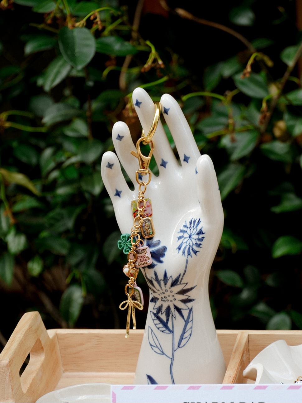 Decorative hand-shaped jewelry holder with earrings and charms, set against green leafy background, on wooden tray.