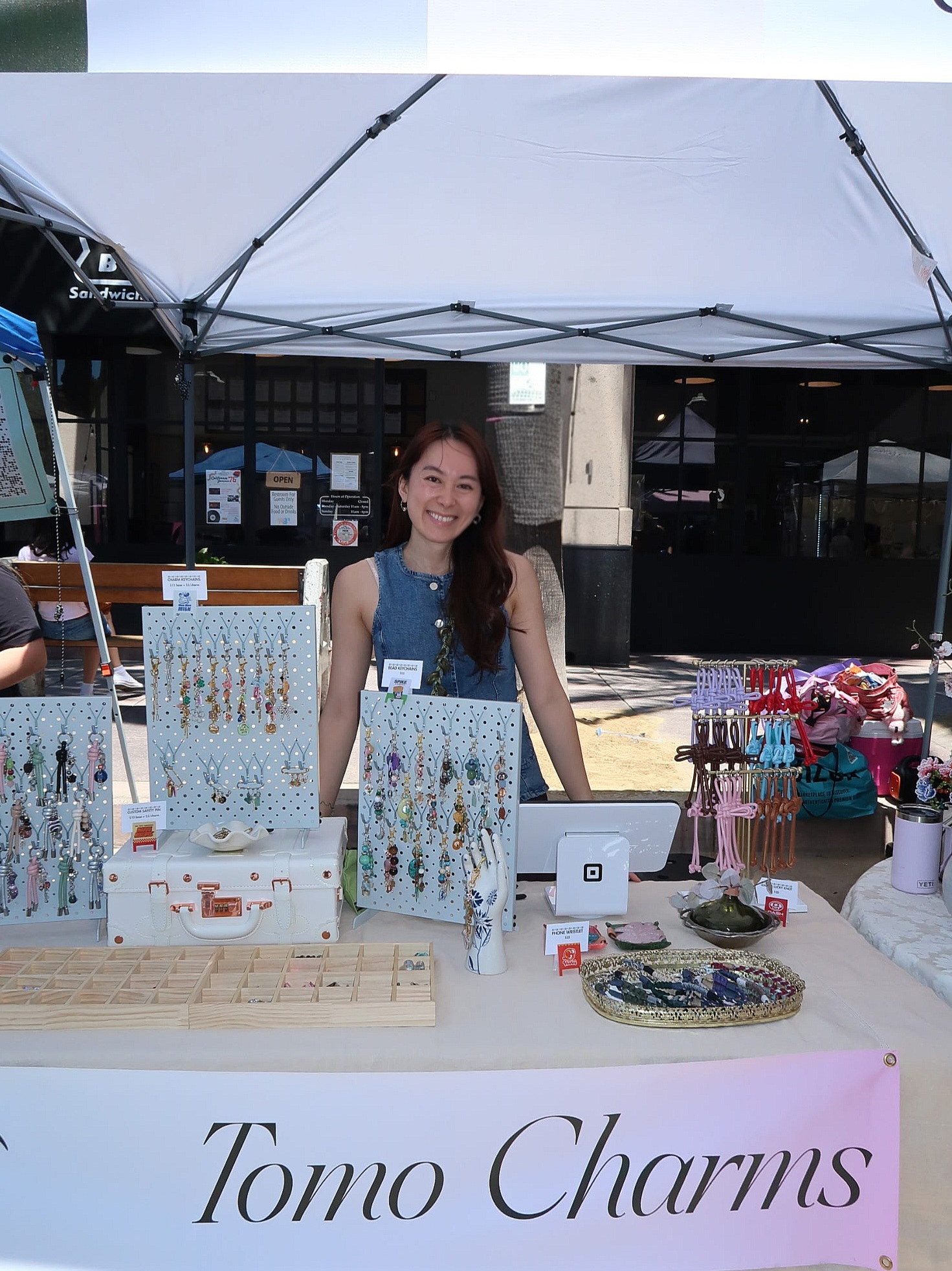Young woman standing behind a table with jewelry and charms at an outdoor market stall under a canopy.