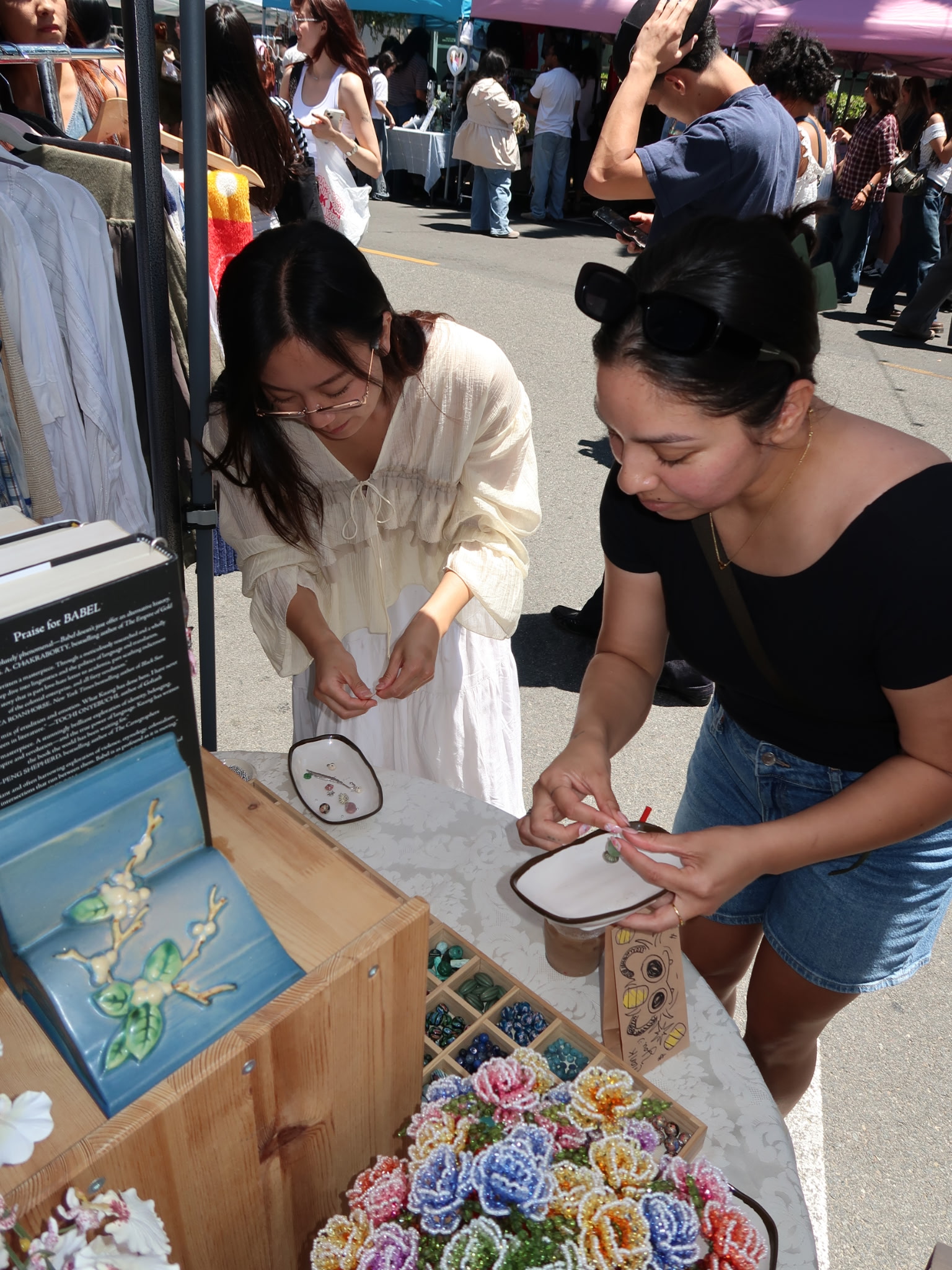 Two women look at jewelry displayed on a table outdoors, with people walking in the background.