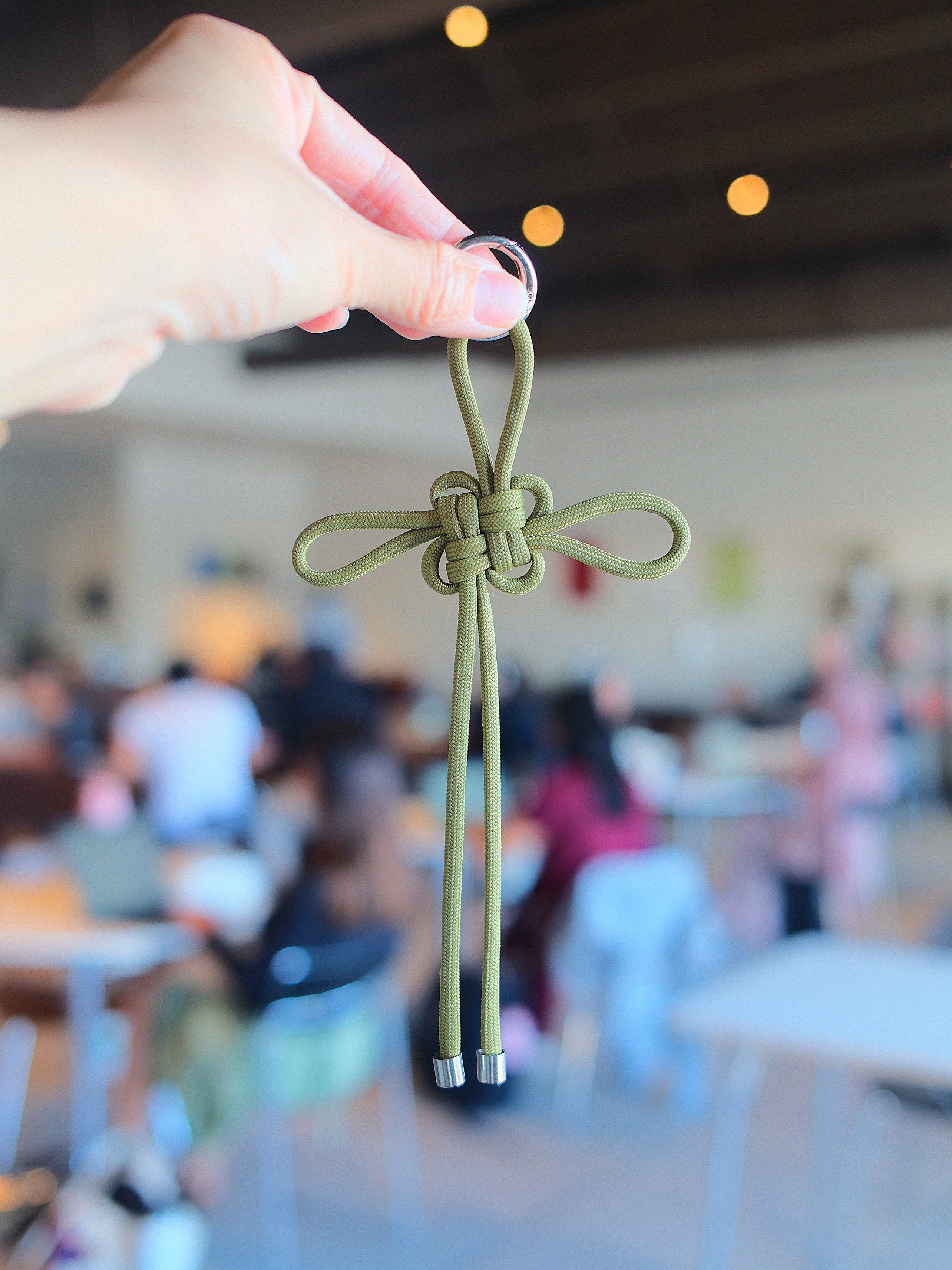 Hand holding a green paracord knot with a blurred indoor background of people and tables.