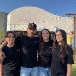 Four people standing outdoors in front of a building with a sign that reads "Sweetably Gelato." They are smiling and wearing black shirts.