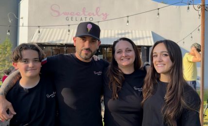 Four people standing outdoors in front of a building with a sign that reads "Sweetably Gelato." They are smiling and wearing black shirts.