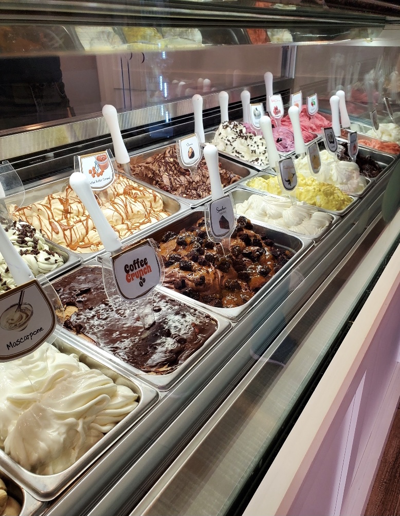 Display case with various flavors of ice cream, each with a white spatula, in a refrigerated showcase.