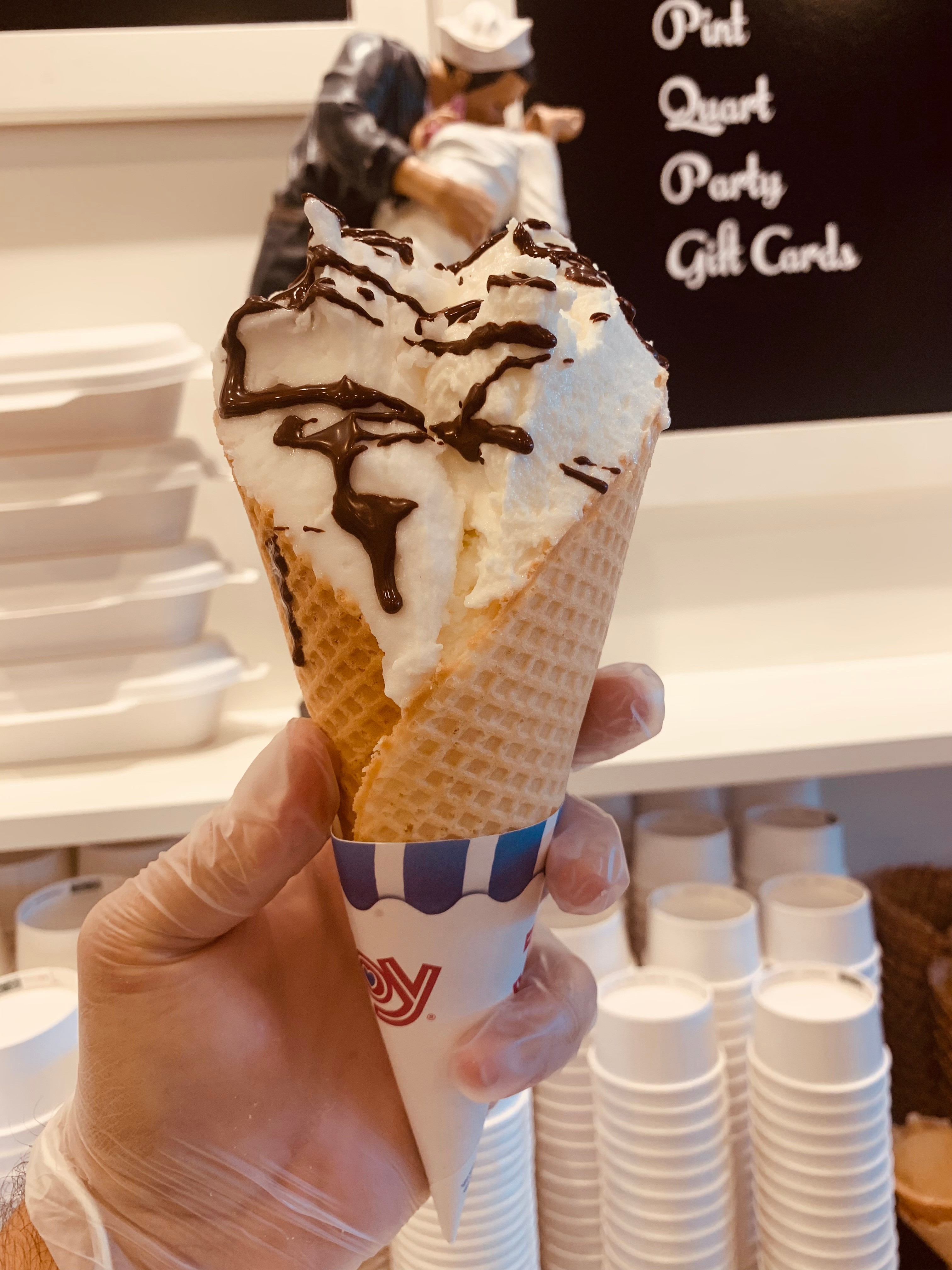 Hand holding an ice cream cone with chocolate drizzle, in front of a blackboard with white text, and stacked cups in background.
