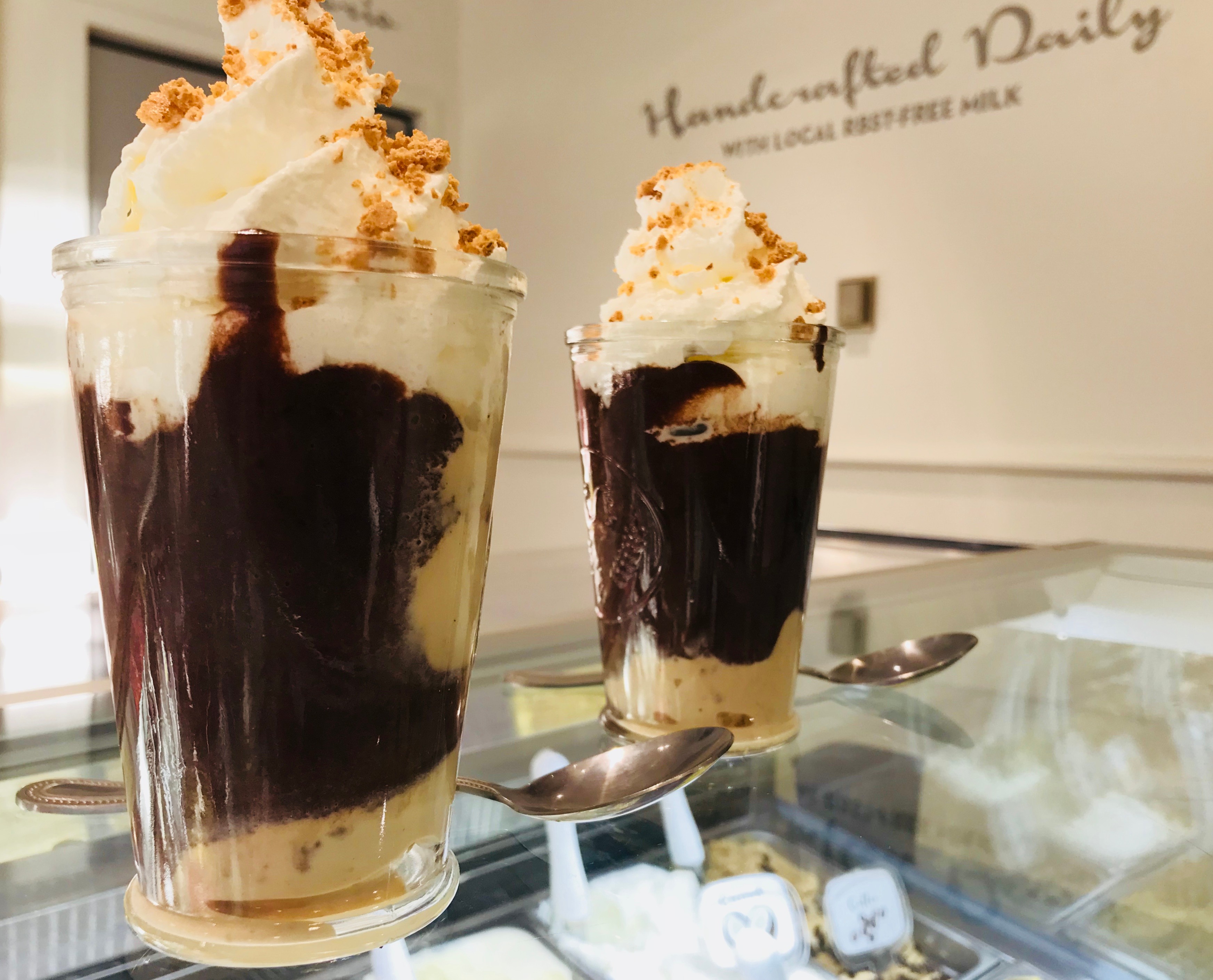 Two tall glasses of layered dessert with whipped cream and chocolate sauce, on a glass surface, with a blurred background.
