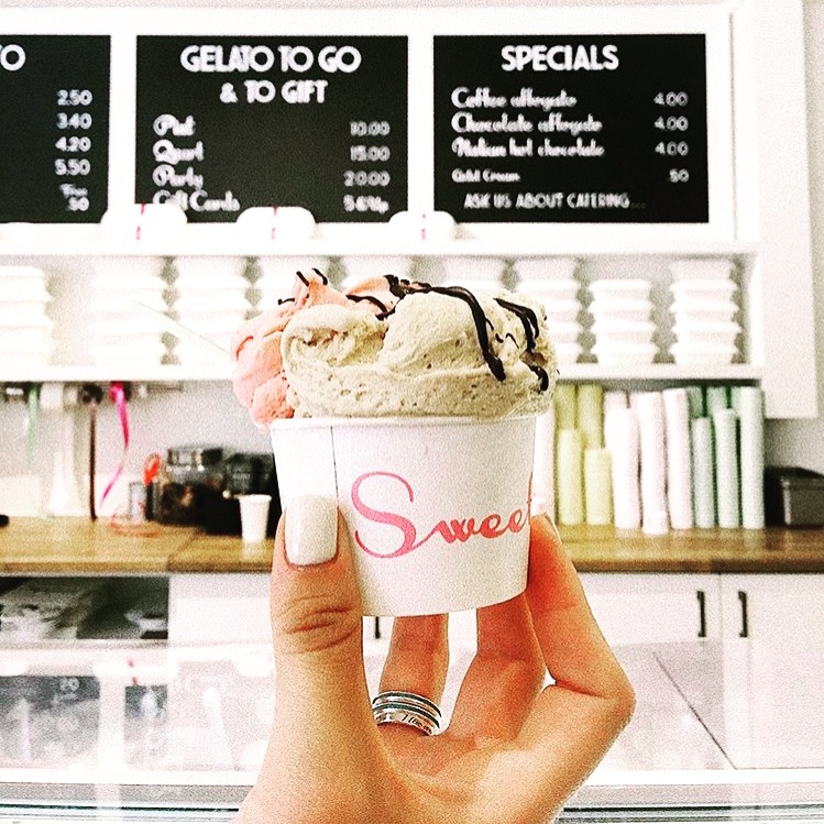 Hand holding a cup of ice cream with three scoops, chocolate drizzle, in a shop with menu boards in the background.
