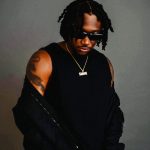 Man with dreadlocks wearing sunglasses, a black sleeveless shirt, and a gold chain, standing against a gray background.