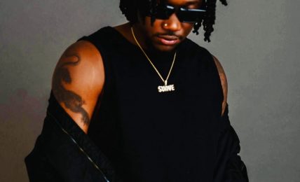 Man with dreadlocks wearing sunglasses, a black sleeveless shirt, and a gold chain, standing against a gray background.