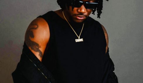 Man with dreadlocks wearing sunglasses, a black sleeveless shirt, and a gold chain, standing against a gray background.