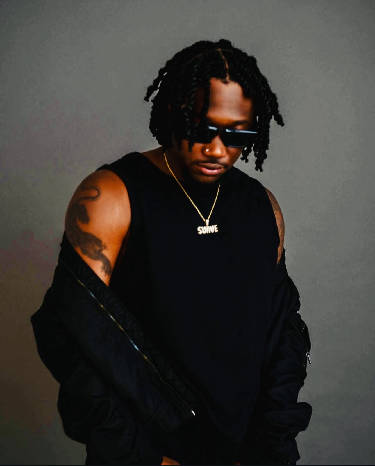 Man with dreadlocks wearing sunglasses, a black sleeveless shirt, and a gold chain, standing against a gray background.