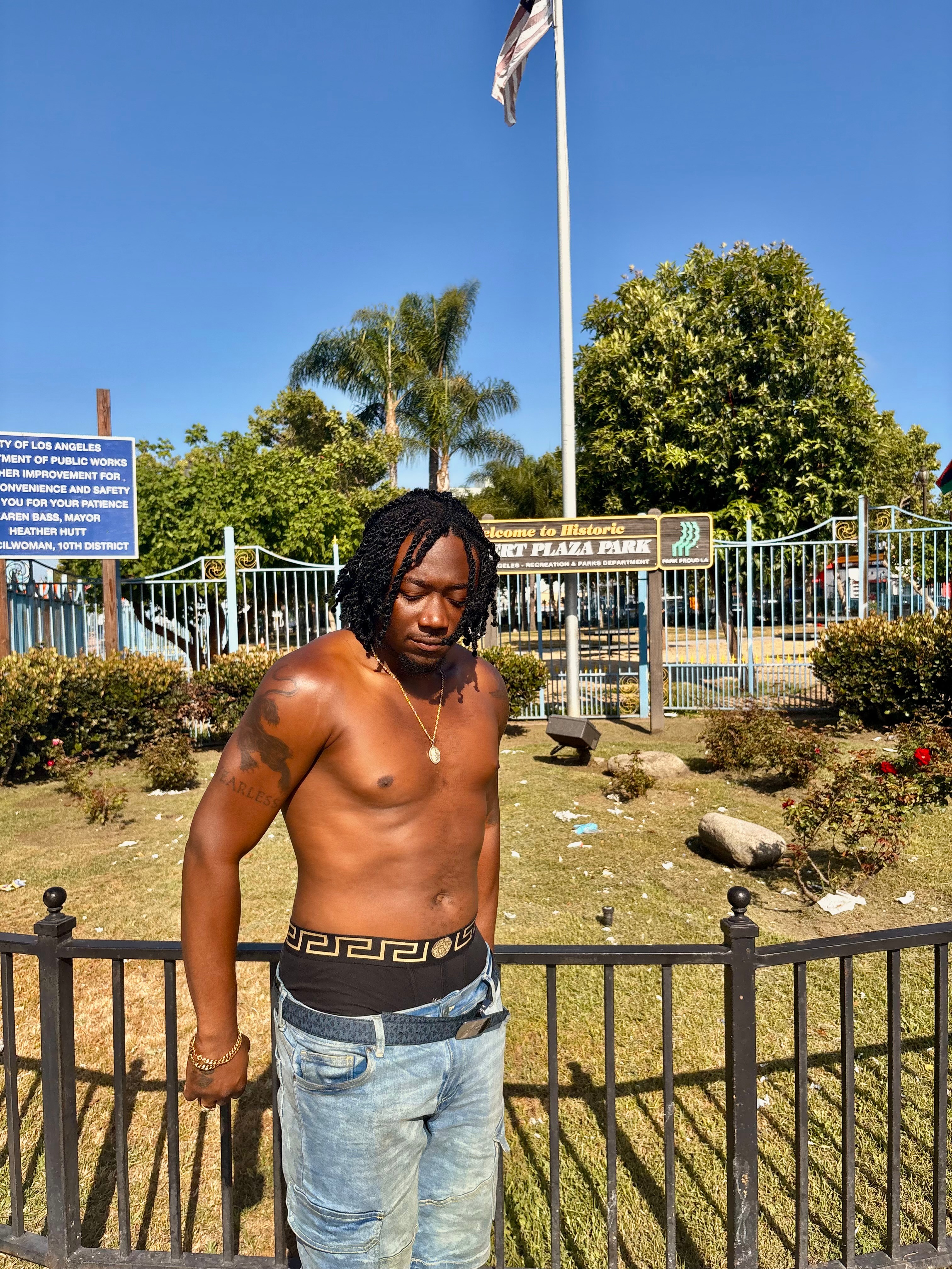 Young man with dreadlocks shirtless standing outdoors in front of a fence and trees, sunny day, blue sky.