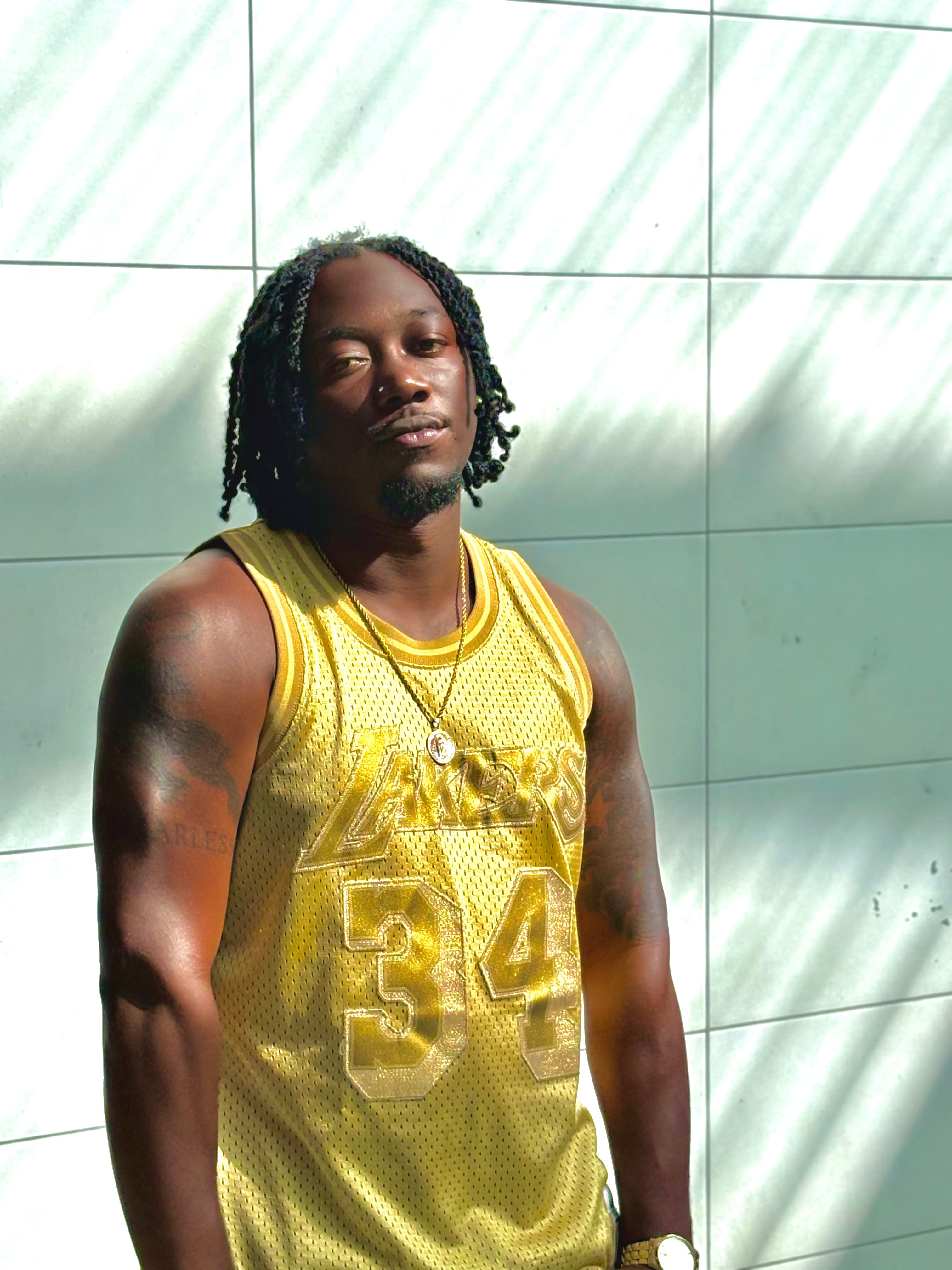 Young man with dreadlocks wearing a yellow sports jersey, standing against a tiled wall with sunlight and shadows.