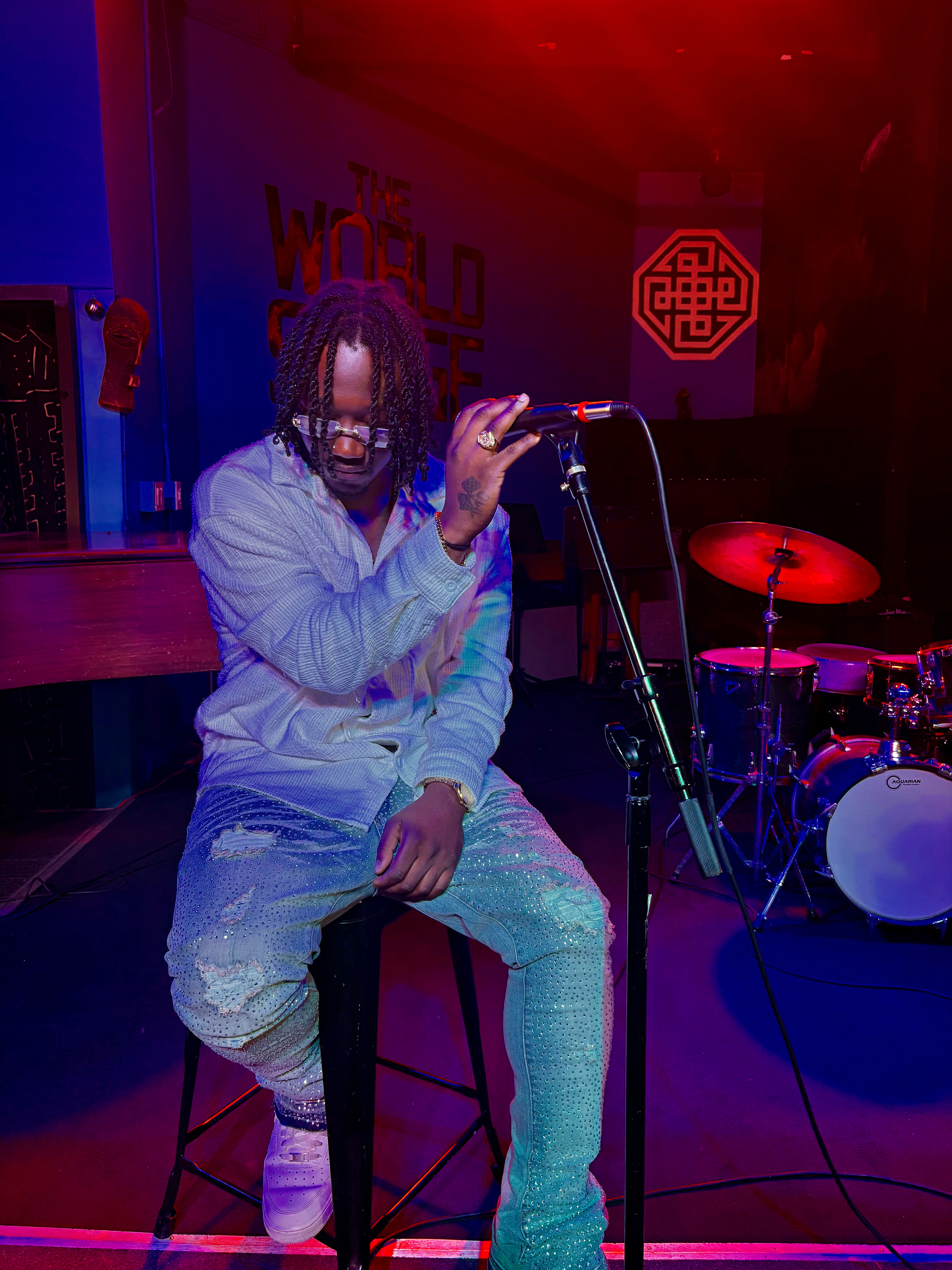 Person sitting on a stool holding a microphone on stage with drums and a sign in the background, dimly lit with red and blue lights.