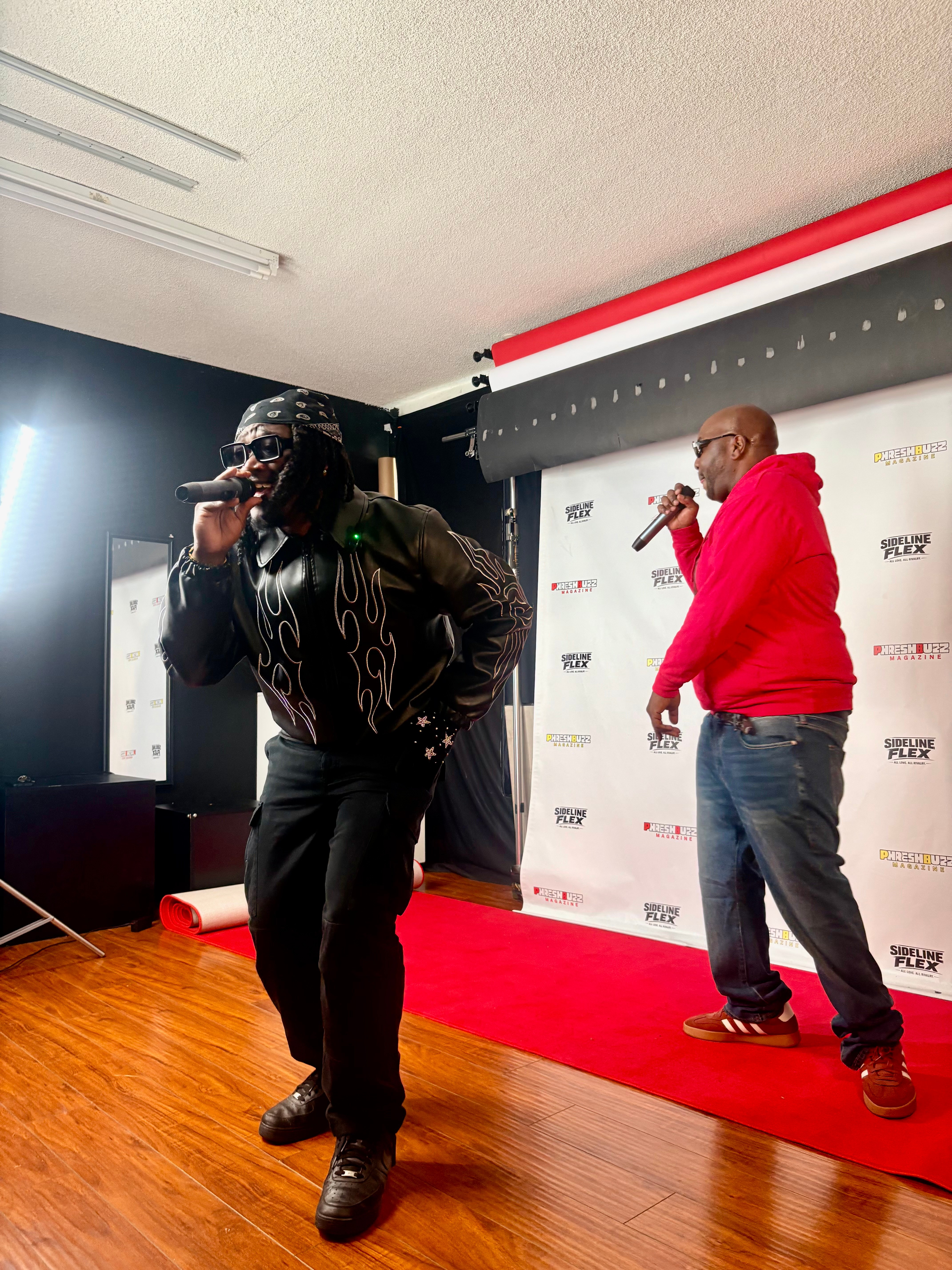 Two men performing on stage with a red carpet, one in a black outfit and the other in a red hoodie, holding microphones.