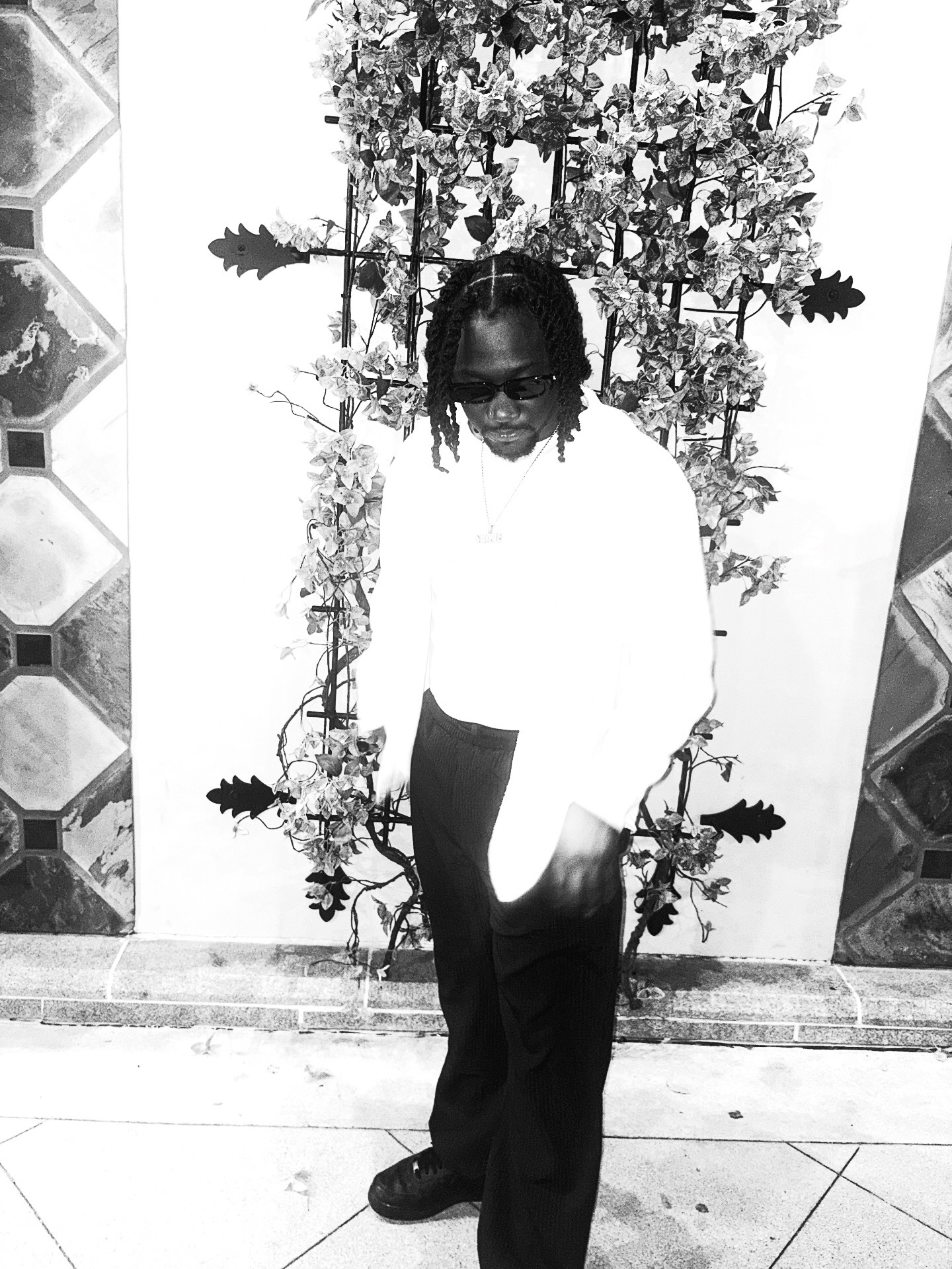 Person standing in front of a floral display, wearing dark sunglasses, a white top, and dark pants.