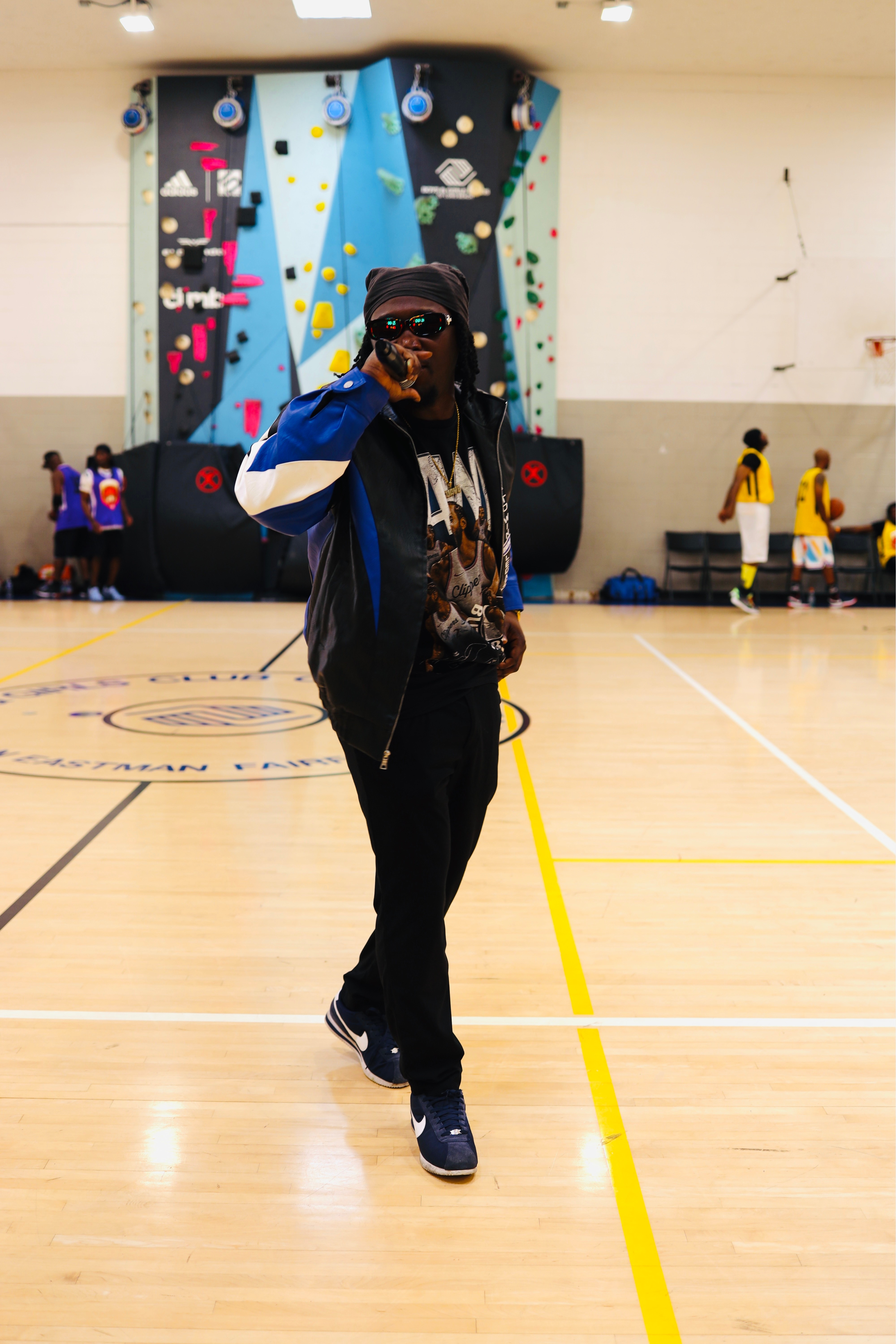 Person standing in a gymnasium with a climbing wall in the background, wearing a black jacket, pants, and sneakers.