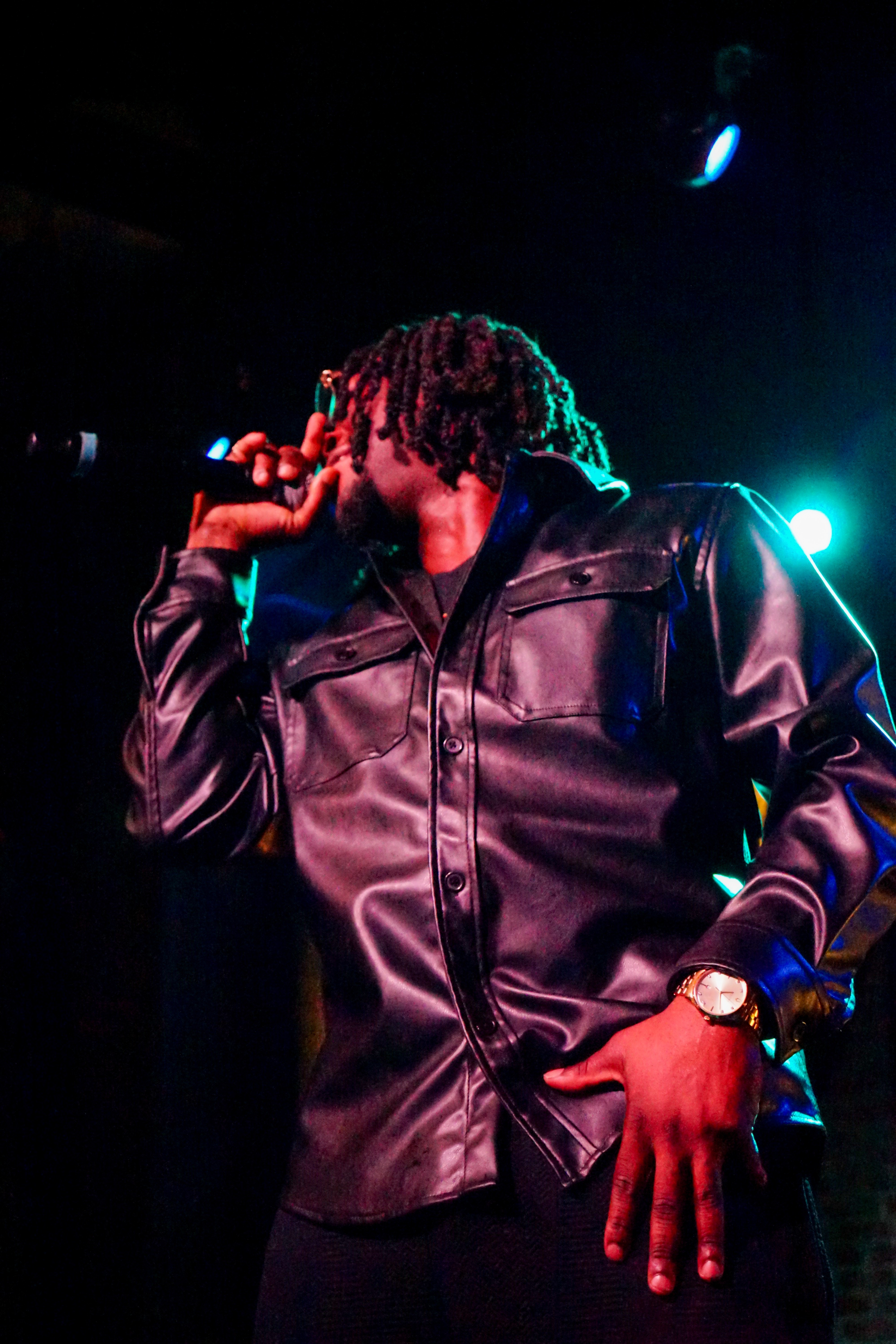 Person with dreadlocks wearing a leather jacket, holding a microphone, performing on stage with blue and green lighting