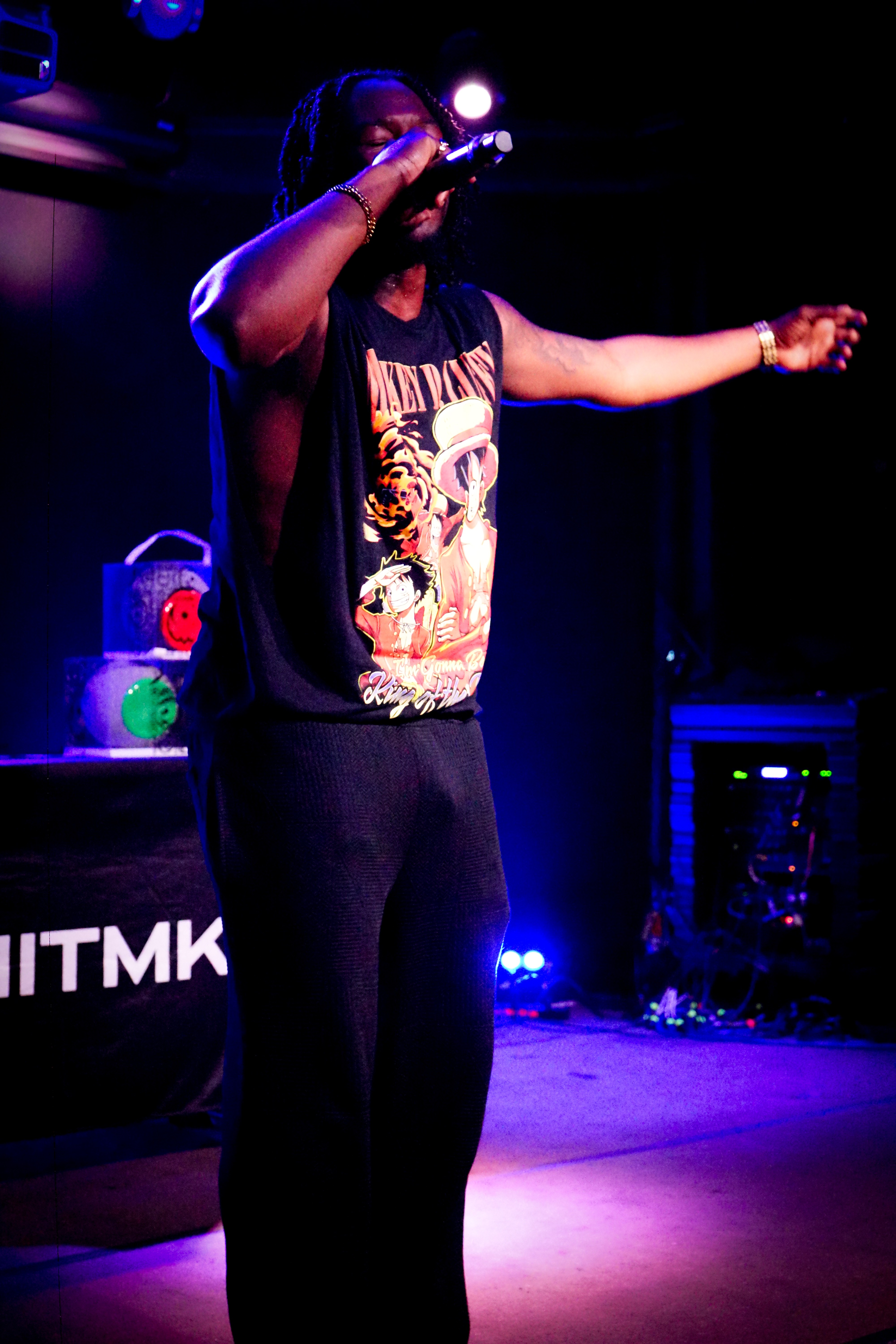 Person singing into a microphone on stage with colorful lights, wearing a graphic sleeveless top and black pants.