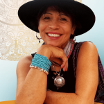 Smiling woman wearing a wide-brimmed hat, colorful bracelets, and a necklace, resting her chin on her hand.