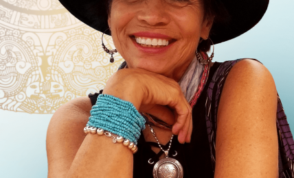 Smiling woman wearing a wide-brimmed hat, colorful bracelets, and a necklace, resting her chin on her hand.