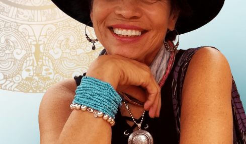 Smiling woman wearing a wide-brimmed hat, colorful bracelets, and a necklace, resting her chin on her hand.